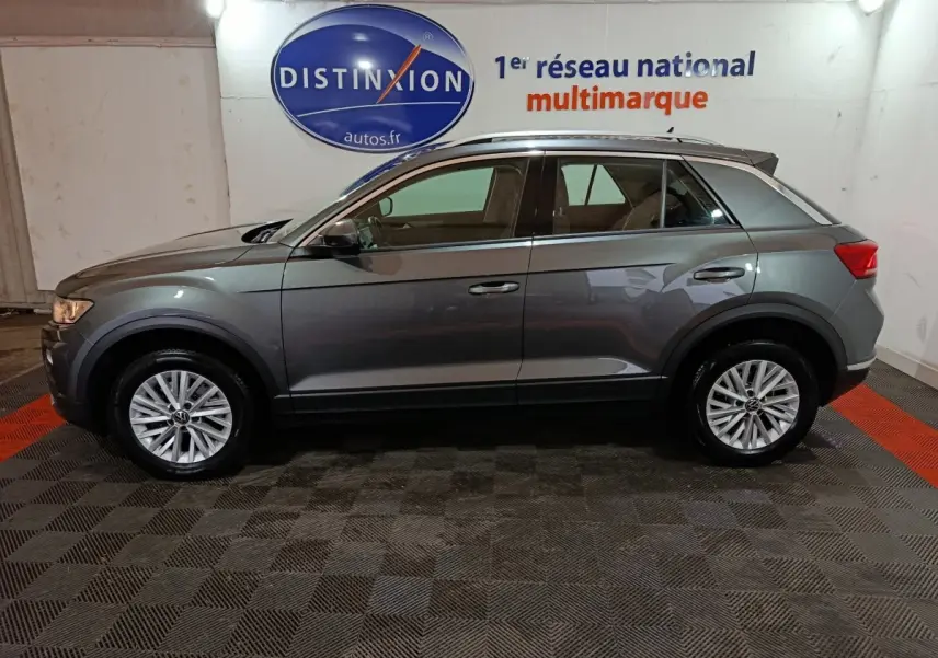 Vue latérale droite d'un Volkswagen T-Roc gris foncé 2021 dans un showroom avec logo Distinxion en arrière-plan.