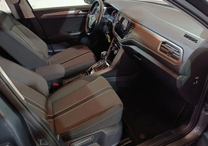 Vue intérieure côté conducteur du Volkswagen T-Roc gris foncé, mettant en valeur les sièges tissu et le tableau de bord moderne avec écran tactile.