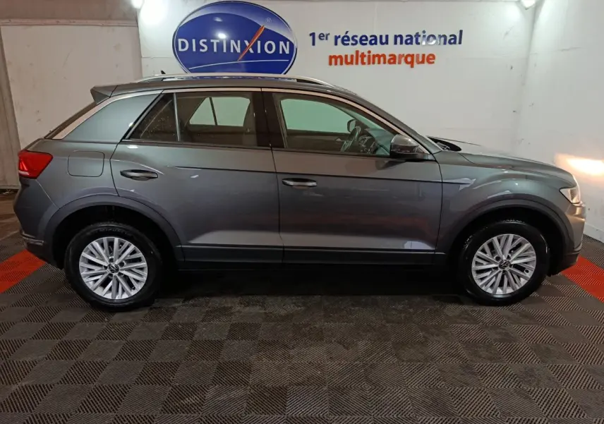 Vue de profil côté gauche d'un Volkswagen T-Roc gris foncé 2021 dans un showroom avec logo Distinxion en arrière-plan.
