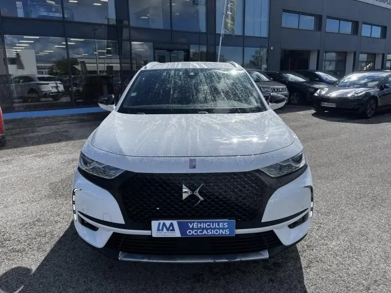 Vue avant d'un DS7 Crossback blanc 2019 avec calandre noire et logo DS, stationné devant un bâtiment vitré.