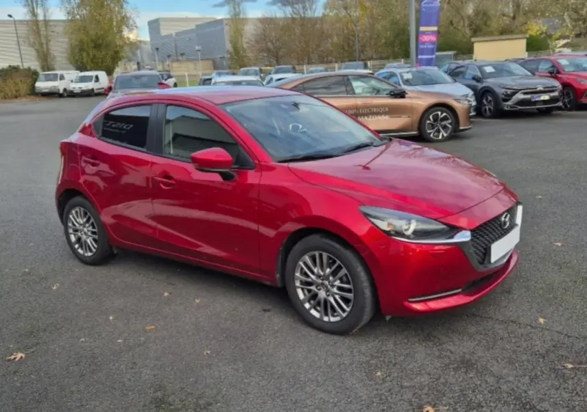 Vue 3/4 avant gauche d'une Mazda2 rouge brillante avec jantes aluminium et vitres surteintées sur parking.