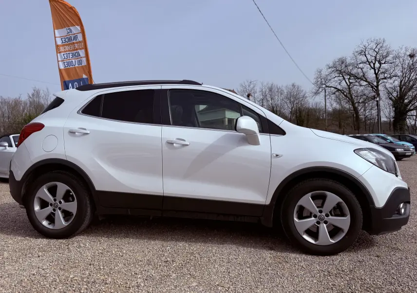 Vue latérale droite d’un Opel Mokka X blanc 5 portes garé sur un sol gravillonné avec un drapeau orange en arrière-plan.
