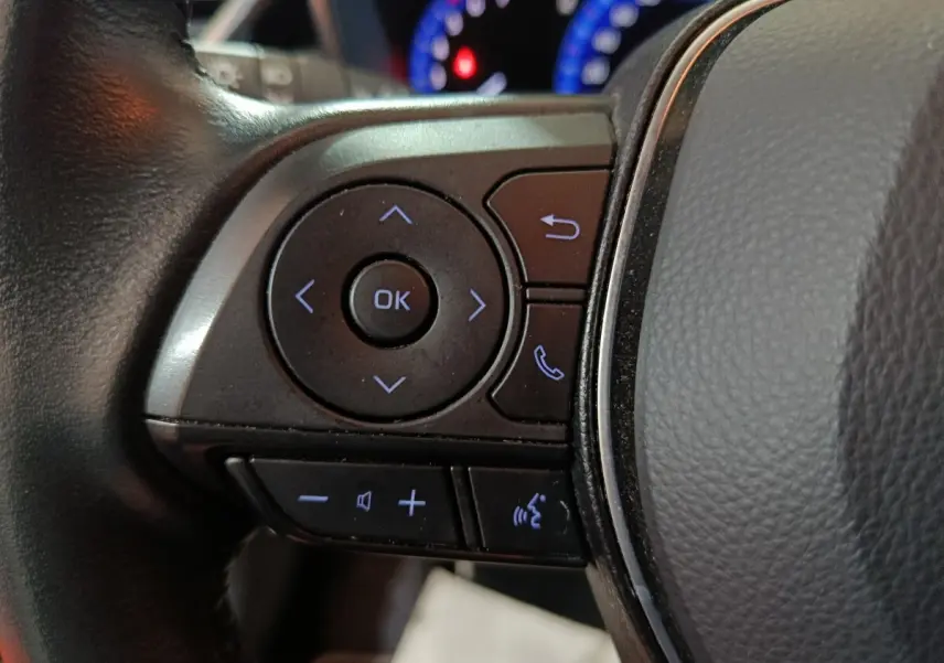 Gros plan sur les commandes au volant côté gauche de la Toyota Corolla hybride blanche, avec boutons multifonctions rétroéclairés.