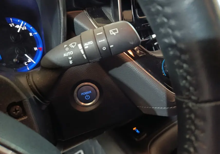 Gros plan intérieur sur le bouton start/stop bleu et la commande d’essuie-glace à gauche du volant cuir d’une Toyota Corolla hybride.