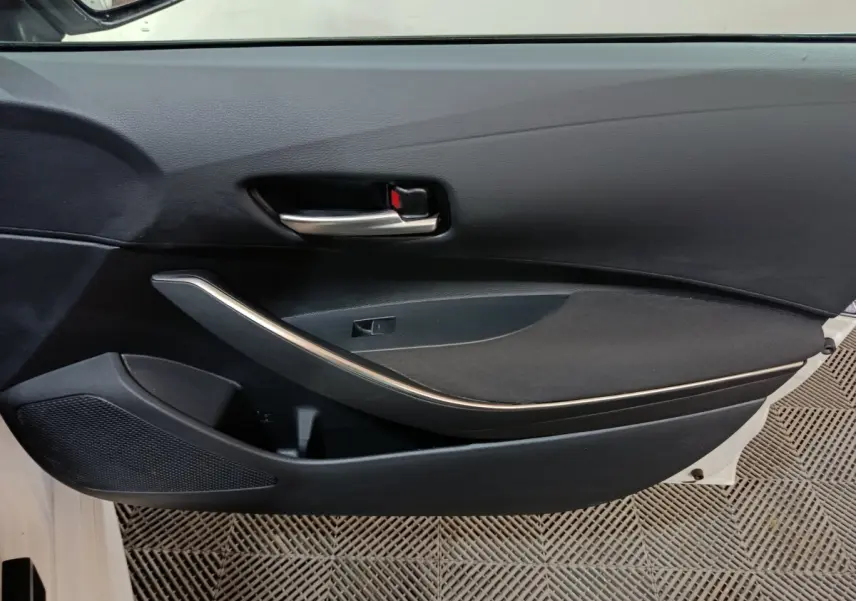 Vue intérieure côté gauche de la porte avant noire de la Toyota Corolla Hybride blanche, avec poignée argentée et haut-parleur intégré.