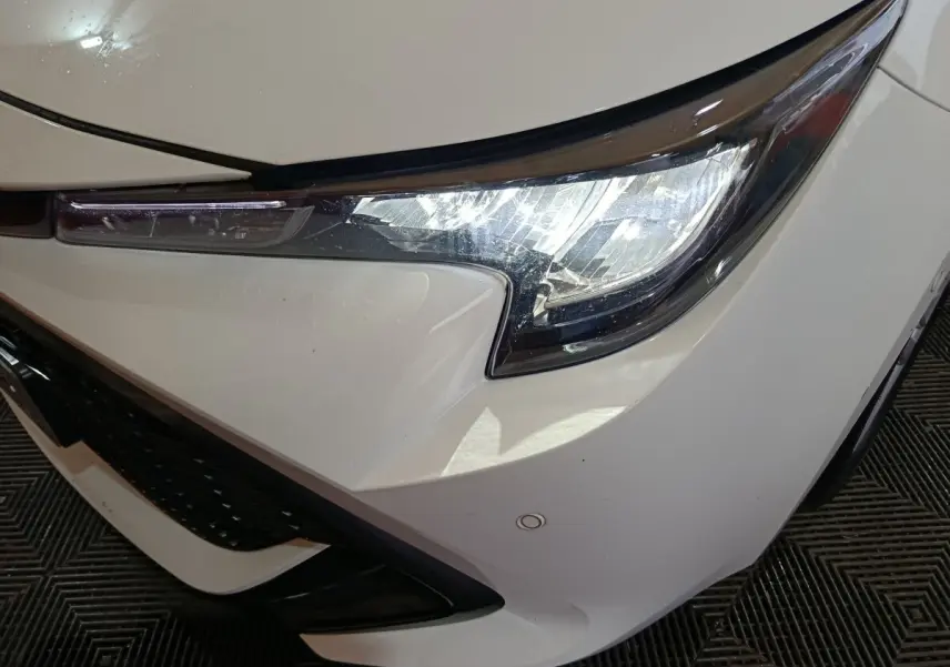 Gros plan sur le phare avant droit blanc d'une Toyota Corolla hybride 2022 avec calandre noire et capteur de stationnement visible.