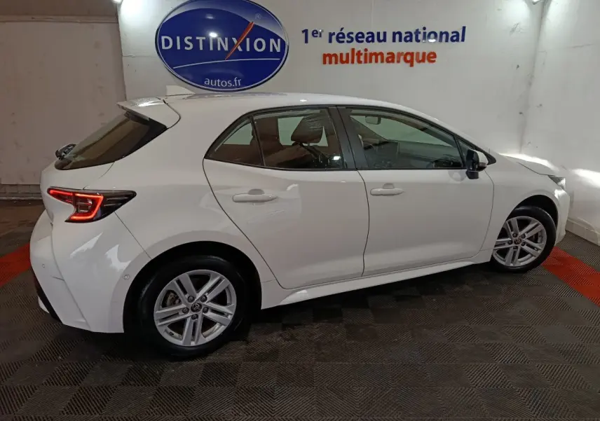 Toyota Corolla hybride blanche vue de profil droit en intérieur avec feux arrière LED allumés et jantes alliage.