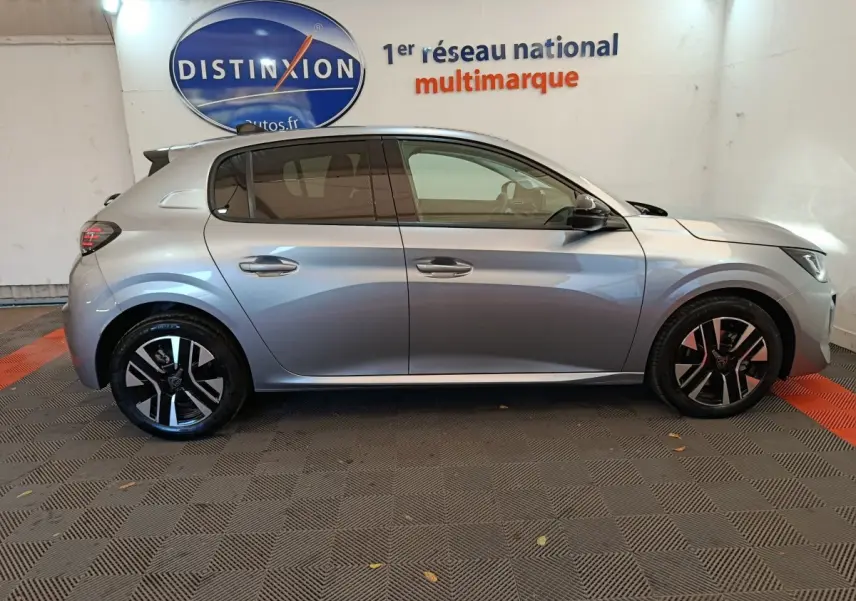 Profil côté droit d'une Peugeot 208 gris clair 2025 garée en intérieur avec jantes noires et logo Distinxion en arrière-plan.