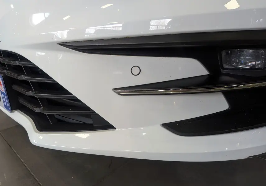 Gros plan sur le pare-chocs avant droit blanc glacier d’une Renault Mégane IV Berline avec radar de stationnement et antibrouillard.
