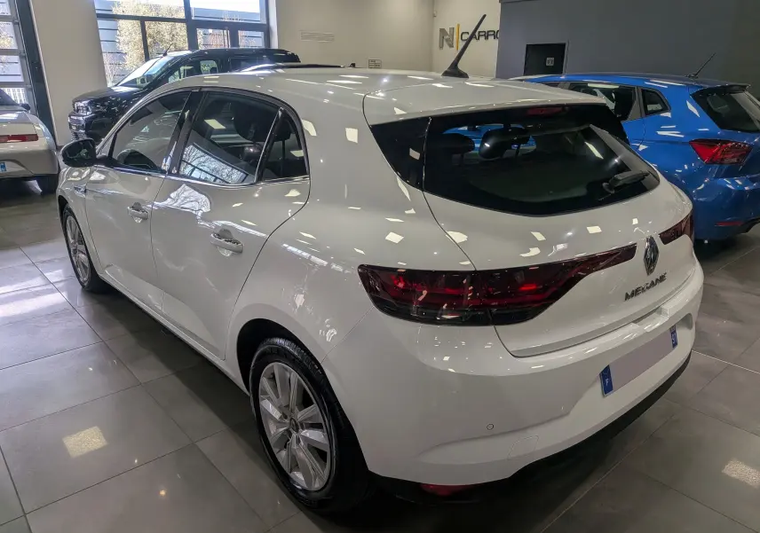 Vue 3/4 arrière droite d’une Renault Mégane IV Berline blanche Glacier, avec feux arrière LED et jantes alliage visibles en intérieur showroom.