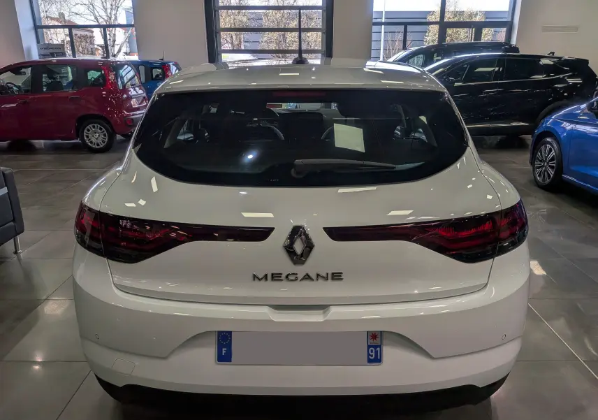 Vue arrière d'une Renault Mégane IV Berline blanc glacier, avec feux arrière allongés et logo central visible en intérieur showroom.