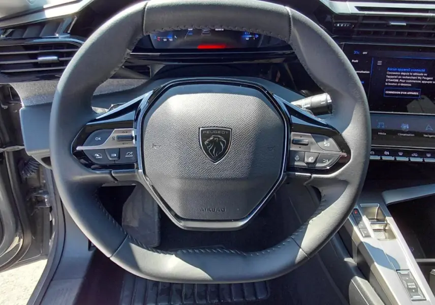 Vue intérieure centrée sur le volant cuir noir de la Peugeot 308 SW Hybrid 2025 avec tableau de bord numérique et écran tactile.