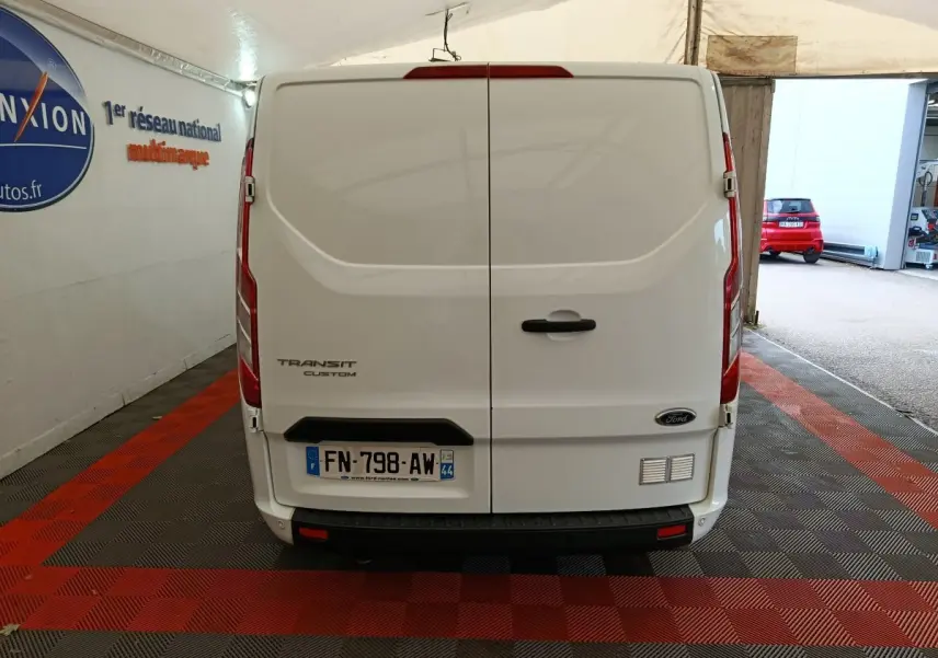 Vue arrière du fourgon blanc Ford Transit Custom, portes fermées, dans un garage avec sol à damier rouge et noir.