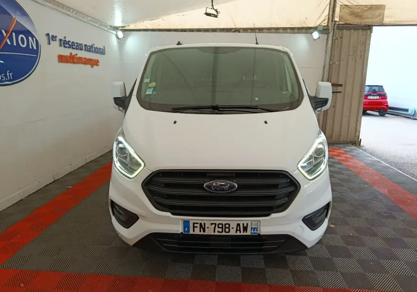 Vue avant d'un Ford Transit Custom blanc avec phares allumés dans un espace couvert.