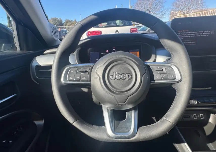 Vue intérieure centrée sur le volant Jeep noir avec commandes intégrées, tableau de bord et écran tactile visibles.