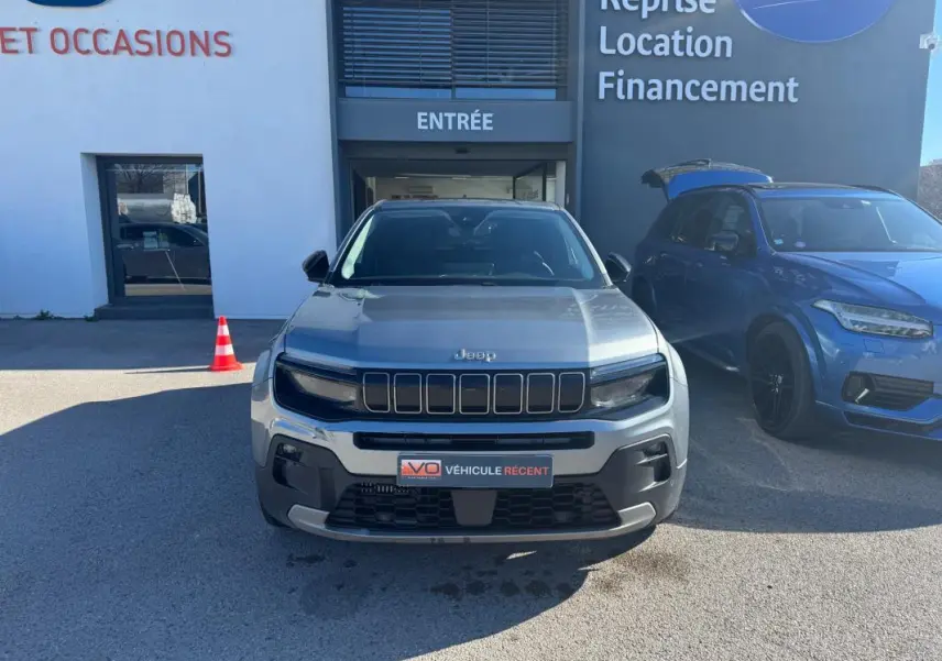 Vue avant d'un Jeep Avenger gris 2025 avec calandre noire et phares LED, stationné devant un bâtiment commercial.
