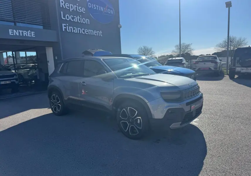 Jeep Avenger gris en 3/4 avant droit, stationné devant un bâtiment avec jantes alliage 18 pouces visibles.