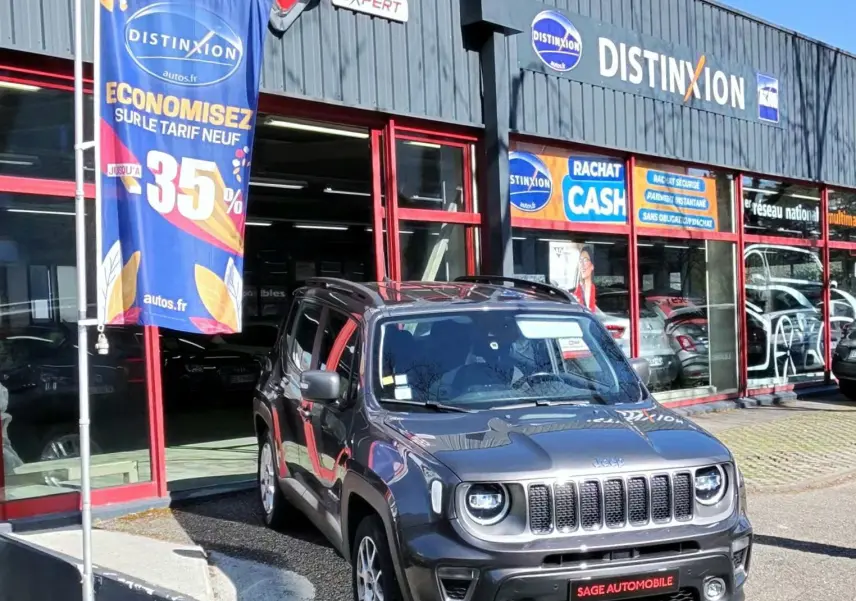Jeep Renegade Granite Crystal métallisé en 3/4 avant droit, stationnée devant un garage avec enseignes commerciales.