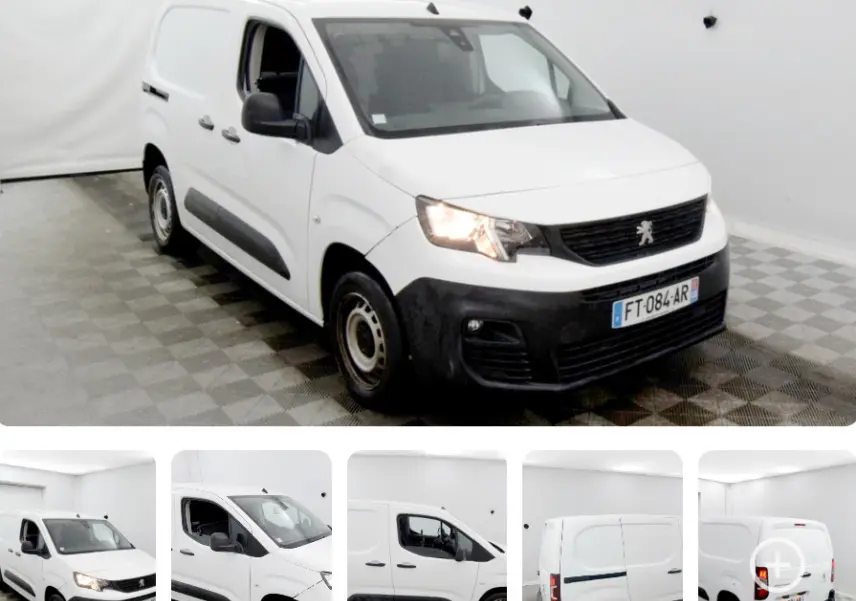 Peugeot Partner blanc vue 3/4 avant droit en intérieur, avec phares allumés et protections noires sur les portes.