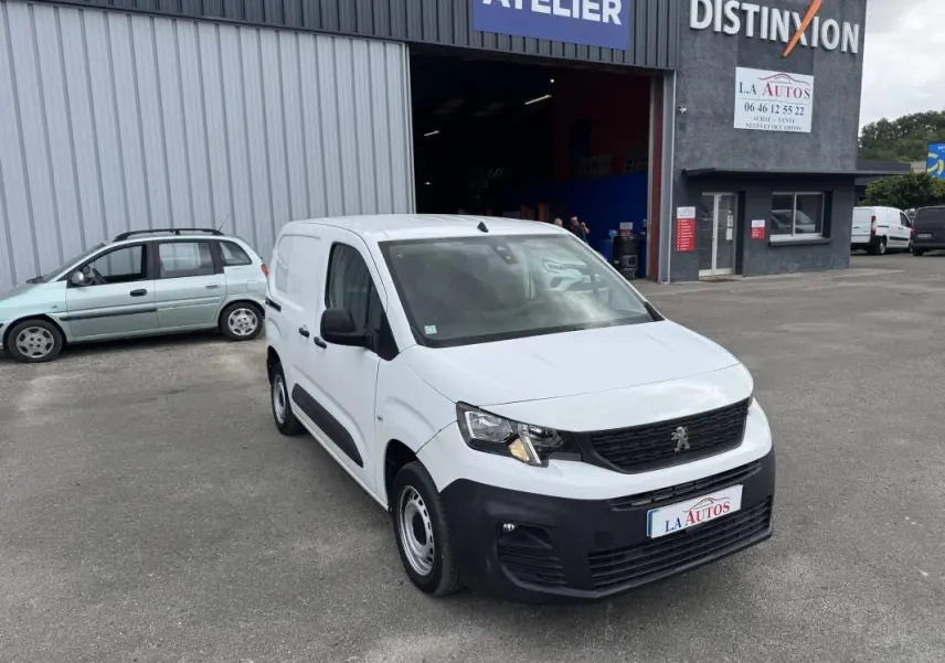 Peugeot Partner blanc vue 3/4 avant droit, avec pare-chocs noir, stationné devant un atelier automobile.