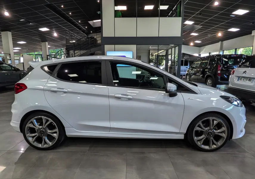 Vue de profil côté gauche d'une Ford Fiesta blanche 1.0 EcoBoost ST Line en intérieur, avec jantes alliage distinctives.