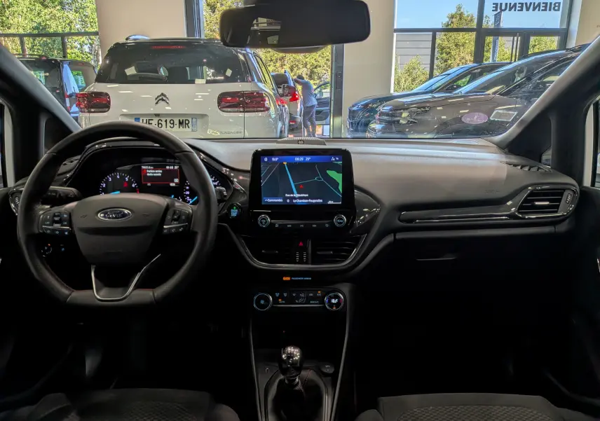 Vue intérieure du tableau de bord et volant de la Ford Fiesta blanche, avec écran tactile central et boîte manuelle visible.