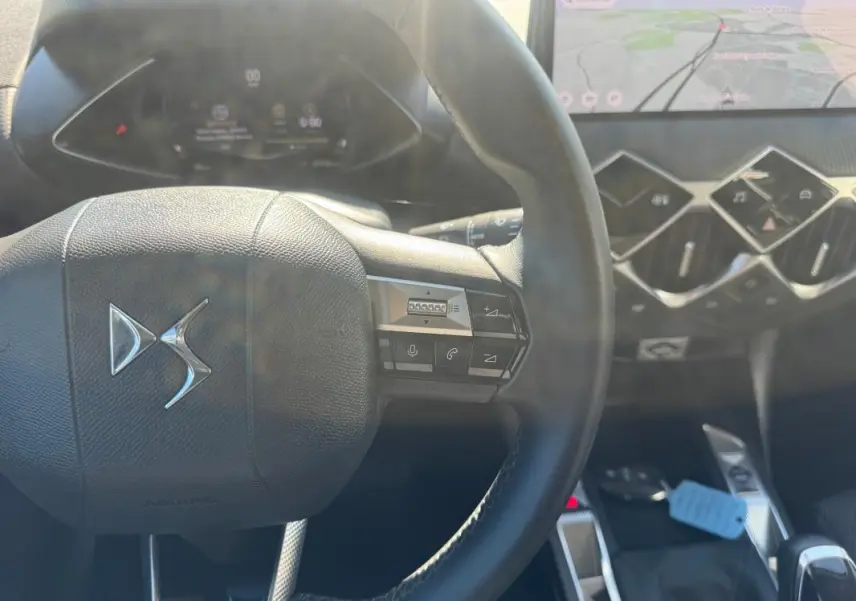 Vue rapprochée du volant DS noir avec commandes intégrées, tableau de bord numérique et écran tactile de navigation en arrière-plan.