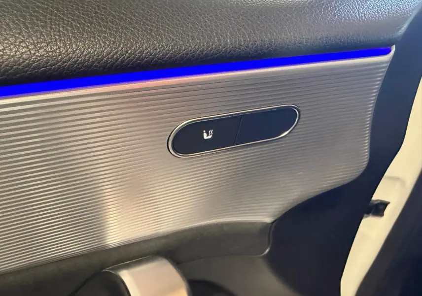 Détail de la porte intérieure côté gauche d’une Mercedes Classe A 220D AMG Line blanche, avec éclairage d’ambiance bleu et bouton de siège chauffant.