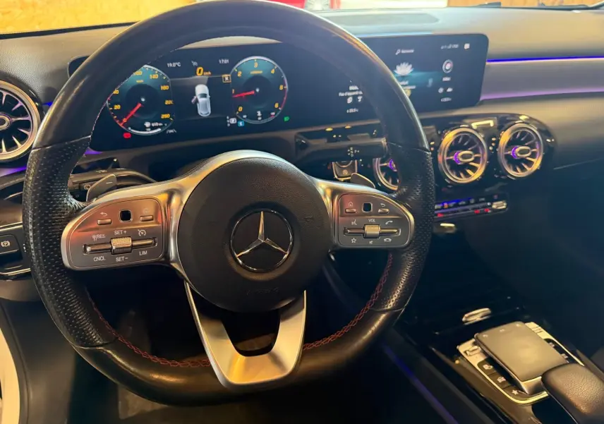 Vue intérieure centrée sur le volant cuir noir de la Mercedes Classe A 220D AMG Line avec tableau de bord numérique et éclairage d'ambiance.