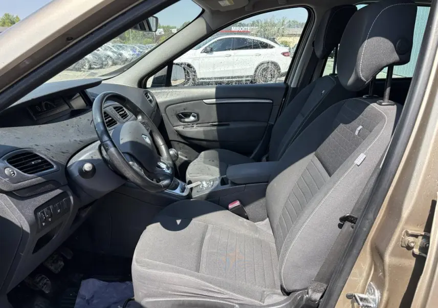 Vue intérieure côté conducteur d’un Renault Scénic III beige 2015, avec volant cuir et accoudoir central visible.