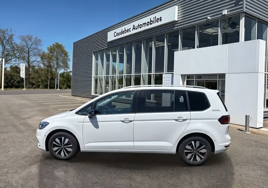 Vue de profil côté gauche d’un Volkswagen Touran blanc 2025 garé devant un bâtiment moderne de concession.