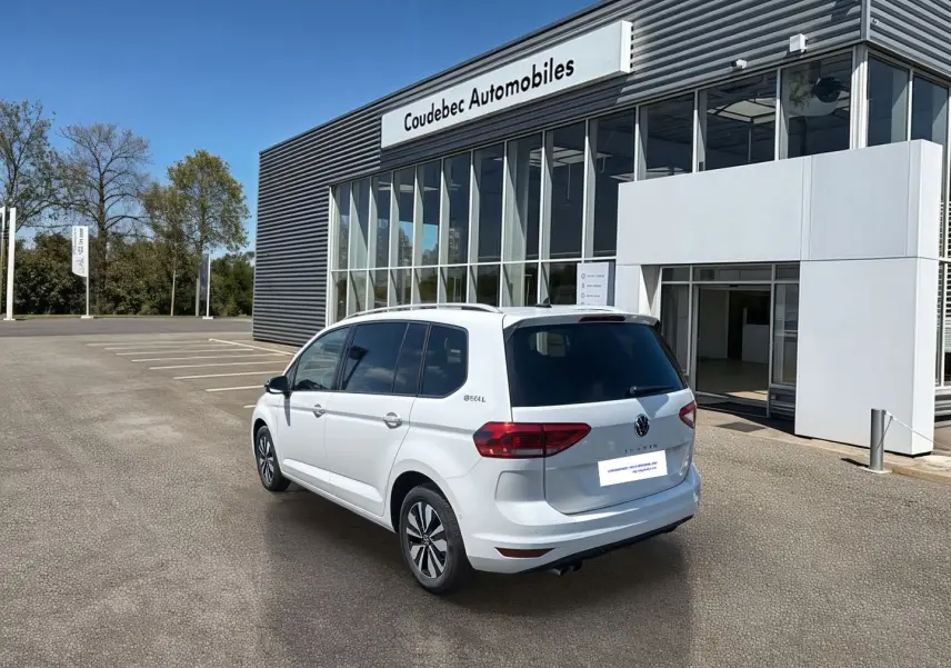 Volkswagen Touran blanc vue 3/4 arrière droit, stationné devant un bâtiment commercial moderne.