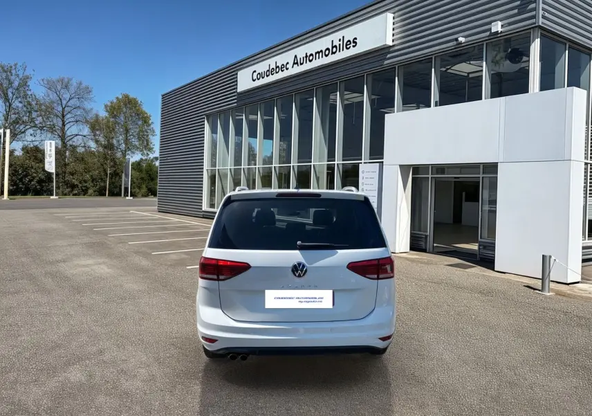 Vue arrière d'un Volkswagen Touran blanc 2025 stationné devant un bâtiment de concession Coudebec Automobiles.