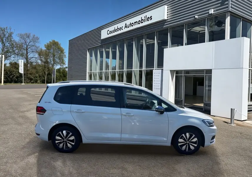 Profil côté gauche d'un Volkswagen Touran blanc 2025 stationné devant un bâtiment moderne de concession.
