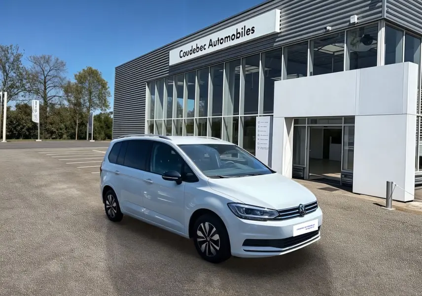 Volkswagen Touran blanc vu en 3/4 avant droit devant un bâtiment moderne de concession automobile.