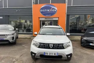 Dacia Duster blanc glacier vu de face, avec calandre noire et barres de toit chromées devant un garage Distinxion.