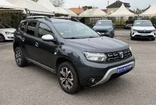 Vue 3/4 avant droit d'un Dacia Duster gris comète 2022 avec barres de toit et jantes alliage distinctives.