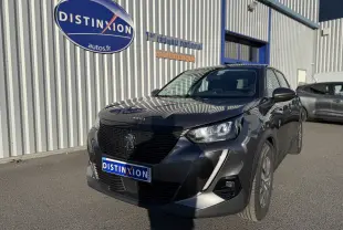 Peugeot 2008 gris 3/4 avant droit, avec calandre noire et phares LED, stationnée devant un bâtiment industriel.