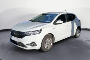Dacia Sandero blanc vue 3/4 avant droit sur parking gravillonné avec plusieurs véhicules en arrière-plan.