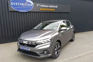 Vue 3/4 avant d'une Dacia Sandero gris schiste 2025 garée devant un mur noir avec logo Distinxion.