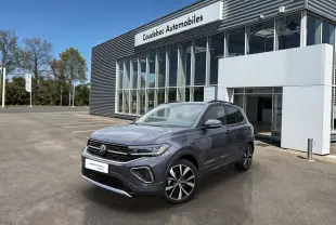 Volkswagen T-Cross gris en 3/4 avant droit, avec jantes alliage noires et grises, devant un bâtiment de concession.