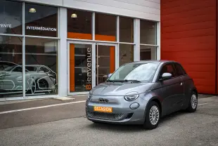 Fiat 500 électrique gris minéral métal vue 3/4 avant avec plaque orange "OCCASION" devant un bâtiment moderne.