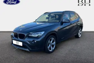BMW X1 sDrive20d 2013 Mineralgrau vue 3/4 avant avec barres de toit noir mat et jantes alu 17 pouces.