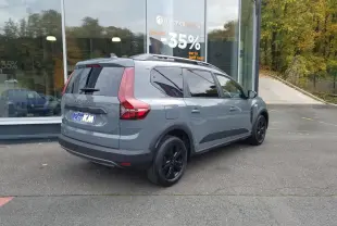 Vue 3/4 arrière droite d'un Dacia Jogger gris urbain avec barres de toit et vitres teintées, stationné devant une vitrine.