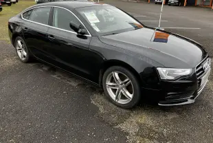 Audi A5 Sportback noir vue 3/4 avant droit, carrosserie brillante avec jantes alliage cinq branches.