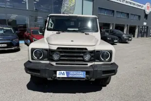 Vue de face de l’INEOS Grenadier beige 2024 avec calandre noire et phares ronds, stationné devant un garage.