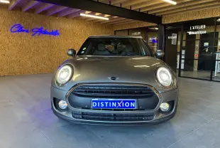 Vue de face d'une MINI Clubman beige 2019 avec phares allumés, calandre noire et plaque Distinxion en intérieur showroom.