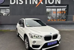 BMW X1 F48 blanc vue 3/4 avant droit avec phares allumés devant un bâtiment Distinxion.