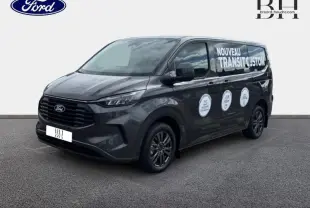 Ford Transit Custom gris métallisé vu en 3/4 avant droit avec marquage publicitaire "Nouveau Transit Custom" sur la porte latérale.