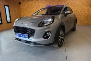 Ford Puma gris en 3/4 avant droit, avec calandre noire et phares allumés dans un garage en bois.